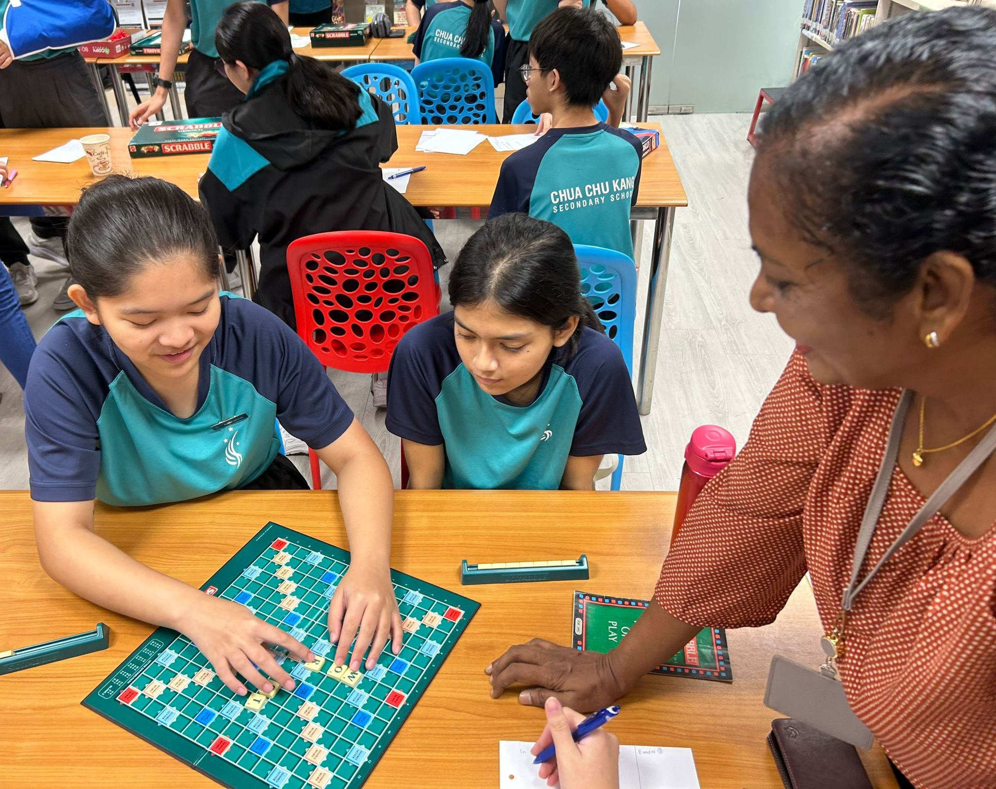 two girls play scrabble as a teacher looks on